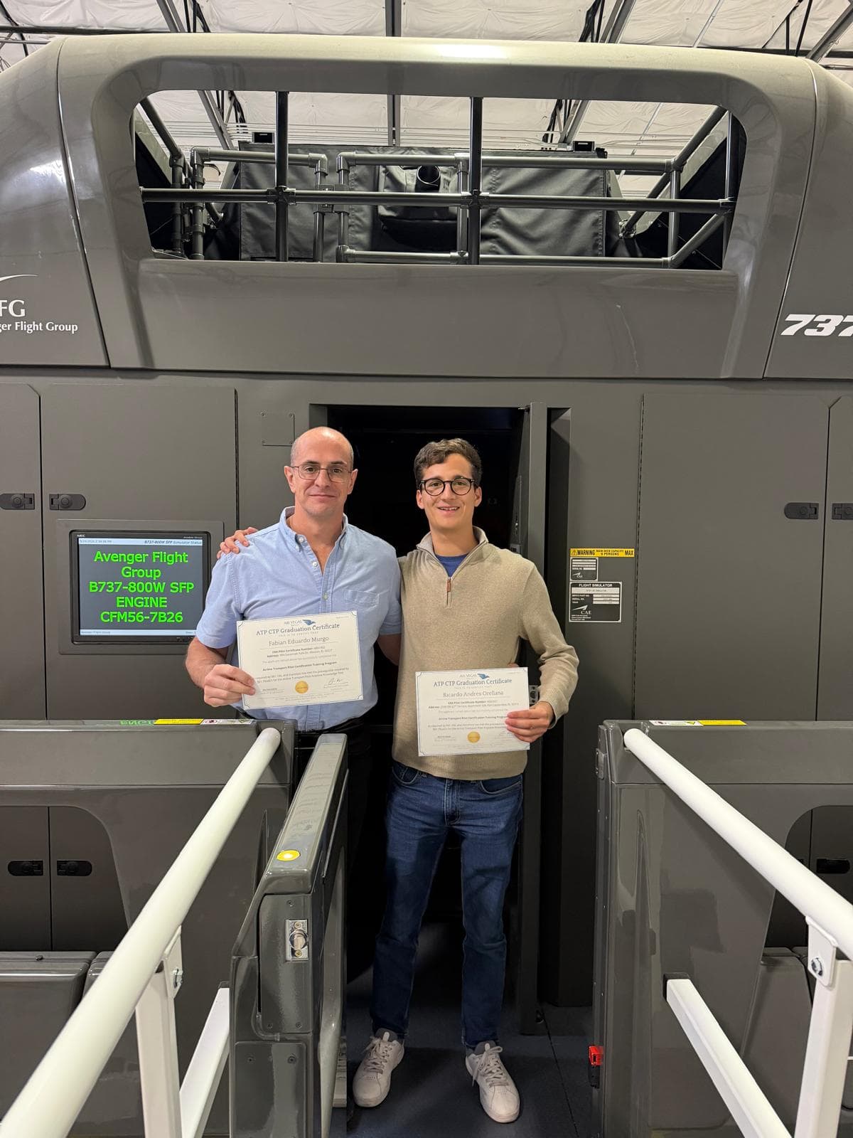 Two AVS graduates holding ATP-CTP certificates in front of the full-motion simulator